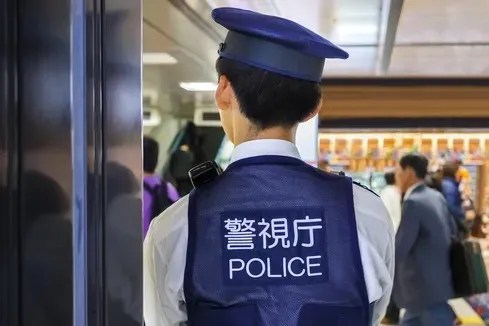 Back of a police officer wearing a dark blue uniform vest with "POLICE" and Japanese characters in white, standing in a public area with people in the background.